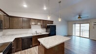 This single-floor residence offers a thoughtfully designed layout that balances functionality and comfort throughout. The heart of the home is an open-concept kitchen outfitted with rich dark wood cabinetry, light marble-style countertops, and black appliances including a refrigerator, range, dishwasher, and over-the-range microwave. A central island provides additional prep space and is illuminated by elegant pendant lighting that hangs from vaulted ceilings, enhancing the sense of openness. The kitchen flows seamlessly into the living area, which features hardwood-style plank flooring, a ceiling fan, and a sliding glass door that opens to a private balcony. A barn-style door adds a modern touch to one of the interior transitions. The bedroom is carpeted for warmth and comfort, equipped with a ceiling fan, a window for natural light, and access to a closet. The bathroom is generously sized, featuring dark wood vanity cabinetry, a light countertop, wood-style tile flooring, and a walk-in shower. A dedicated hallway conveniently houses a stacked in-unit washer and dryer. Baseboard heating is present throughout, and the clean, neutral palette creates a bright and welcoming atmosphere across the entire space.