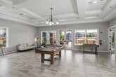 This two-story clubhouse offers an impressive and well-appointed community gathering space spanning two thoughtfully designed floors. The ground floor makes an immediate statement with a grand rotunda entrance hall, defined by soaring ceilings, bold black structural columns, and a striking three-dimensional black-and-white geometric tiled floor. An ornate wrought-iron balcony overhead and a cascading pendant light fixture visible from the upper level add dramatic architectural character to the space.

Adjacent to the entrance, a stylish lounge area features a floor-to-ceiling stone-clad fireplace wall, marble-look tile flooring, and contemporary seating. A bar area with a white quartz countertop, slate-toned cabinetry, and globe pendant lighting provides a refined refreshment station. The ground floor also includes a bright administrative office area with light wood flooring and workspace for staff.

The upper floor, larger in footprint, houses a spacious event and recreation room featuring a coffered ceiling, oversized windows with outdoor views, and large-format tile flooring. A game area with a pool table adds a social focal point. A dedicated conference room with direct balcony access and a connected corridor with elevator access complete the upper level, ensuring functionality throughout.