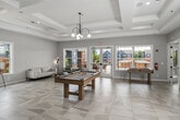 This two-story clubhouse offers an impressive and well-appointed community gathering space spanning two thoughtfully designed floors. The ground floor makes an immediate statement with a grand rotunda entrance hall, defined by soaring ceilings, bold black structural columns, and a striking three-dimensional black-and-white geometric tiled floor. An ornate wrought-iron balcony overhead and a cascading pendant light fixture visible from the upper level add dramatic architectural character to the space.

Adjacent to the entrance, a stylish lounge area features a floor-to-ceiling stone-clad fireplace wall, marble-look tile flooring, and contemporary seating. A bar area with a white quartz countertop, slate-toned cabinetry, and globe pendant lighting provides a refined refreshment station. The ground floor also includes a bright administrative office area with light wood flooring and workspace for staff.

The upper floor, larger in footprint, houses a spacious event and recreation room featuring a coffered ceiling, oversized windows with outdoor views, and large-format tile flooring. A game area with a pool table adds a social focal point. A dedicated conference room with direct balcony access and a connected corridor with elevator access complete the upper level, ensuring functionality throughout.