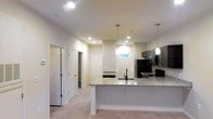 This single-floor residence is designed with an open-concept layout that connects the kitchen to the surrounding living areas. The kitchen serves as a central focal point, featuring a spacious island with light-toned granite countertops that provide ample workspace and a natural gathering area. Dark cabinetry lines the kitchen walls, offering generous storage, while black appliances — including a refrigerator, range, and built-in microwave — add a cohesive, modern aesthetic. Two pendant lights hang above the island, complementing the recessed lighting throughout the space to create a well-lit environment.

A small horizontal window above the countertop near the back wall brings in a touch of natural light, offering a glimpse of greenery outside. The flooring transitions from the kitchen area into carpeted spaces in the adjoining hallways and rooms, softening the overall feel of the interior. Multiple doorways visible from the kitchen suggest well-distributed room access, with hallways leading to additional private spaces. The neutral color palette throughout — warm whites and soft grays — gives the space a clean, understated character that is both practical and welcoming.