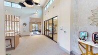 Located at 305 Mezzo Ln in Virginia Beach, VA, this single-floor community clubhouse and leasing center offers a thoughtfully designed, open-concept layout with a generous footprint. Upon entry, soaring double-height ceilings immediately establish a sense of grandeur, complemented by clerestory windows that flood the space with abundant natural light. A striking reception desk crafted from warm wood tones anchors the central area, accented by bold red stools and geometric hexagonal pendant lighting overhead. Vertical wood slat partitions artfully define zones within the open floor plan without interrupting the airy flow of the space. The lounge area features contemporary seating arrangements, including plush sofas and accent chairs, positioned around a linear fireplace and a wall-mounted flat-screen display — ideal for relaxed gatherings. A dining area with modern tables and chairs sits nearby, reinforcing the space's versatility. Floor-to-ceiling glazed walls in the lounge open to sweeping views of an outdoor pool and landscaped amenity area, seamlessly connecting the interior to the outdoors. Black-framed glass partitions enclose a private office suite, adding a refined industrial touch. The kitchen provides additional functionality for community events. Neutral carpeting, light wood flooring, and a warm, sophisticated palette of cream, tan, and wood tones unify the space throughout.