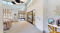 Located at 305 Mezzo Ln in Virginia Beach, VA, this single-floor community clubhouse and leasing center offers a thoughtfully designed, open-concept layout with a generous footprint. Upon entry, soaring double-height ceilings immediately establish a sense of grandeur, complemented by clerestory windows that flood the space with abundant natural light. A striking reception desk crafted from warm wood tones anchors the central area, accented by bold red stools and geometric hexagonal pendant lighting overhead. Vertical wood slat partitions artfully define zones within the open floor plan without interrupting the airy flow of the space. The lounge area features contemporary seating arrangements, including plush sofas and accent chairs, positioned around a linear fireplace and a wall-mounted flat-screen display — ideal for relaxed gatherings. A dining area with modern tables and chairs sits nearby, reinforcing the space's versatility. Floor-to-ceiling glazed walls in the lounge open to sweeping views of an outdoor pool and landscaped amenity area, seamlessly connecting the interior to the outdoors. Black-framed glass partitions enclose a private office suite, adding a refined industrial touch. The kitchen provides additional functionality for community events. Neutral carpeting, light wood flooring, and a warm, sophisticated palette of cream, tan, and wood tones unify the space throughout.