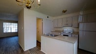 This two-story residence offers a practical and well-organized layout across both levels. The kitchen serves as a central feature of the main floor, designed with a peninsula-style countertop that provides additional workspace and creates a natural division between the kitchen and adjoining areas. Light-toned cabinetry lines the walls, offering ample storage, while a white electric range and an under-cabinet range hood provide essential cooking functionality. A double-basin sink is integrated into the peninsula, adding convenience to meal preparation and cleanup. The countertops feature a neutral, speckled laminate finish that complements the overall color palette of the space. Overhead, a brass chandelier adds a classic decorative touch, accompanied by a flush-mount ceiling fixture that ensures the kitchen remains well-lit. Hardwood-style flooring runs throughout the visible areas, contributing warmth and a cohesive aesthetic. Doorways visible from the kitchen hint at additional rooms and storage spaces, including what appears to be a closet and a hallway leading further into the home. The walls are finished in a soft, neutral tone, lending the space a clean and understated character throughout both floors.