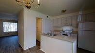 This two-story residence offers a practical and well-organized layout across both levels. The kitchen serves as a central feature of the main floor, designed with a peninsula-style countertop that provides additional workspace and creates a natural division between the kitchen and adjoining areas. Light-toned cabinetry lines the walls, offering ample storage, while a white electric range and an under-cabinet range hood provide essential cooking functionality. A double-basin sink is integrated into the peninsula, adding convenience to meal preparation and cleanup. The countertops feature a neutral, speckled laminate finish that complements the overall color palette of the space. Overhead, a brass chandelier adds a classic decorative touch, accompanied by a flush-mount ceiling fixture that ensures the kitchen remains well-lit. Hardwood-style flooring runs throughout the visible areas, contributing warmth and a cohesive aesthetic. Doorways visible from the kitchen hint at additional rooms and storage spaces, including what appears to be a closet and a hallway leading further into the home. The walls are finished in a soft, neutral tone, lending the space a clean and understated character throughout both floors.