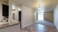 This single-floor residence offers a thoughtfully designed open layout that seamlessly connects the kitchen and living areas. The kitchen features a granite-topped breakfast bar, providing both a functional workspace and a casual dining option. Hardwood-style flooring extends throughout the main living spaces, lending warmth and a cohesive aesthetic to the interior.

A partial wall divides the kitchen from the living area, creating a sense of defined space while maintaining an open feel. The living area is bathed in natural light, thanks to large windows that draw the eye toward the outdoors. An elegant orb-style chandelier serves as a focal point overhead, adding a modern yet classic decorative touch to the space.

The neutral, warm-toned walls throughout complement the light-colored finishes, creating a bright and inviting ambiance. A hallway visible from the main area leads to additional rooms, suggesting a practical and well-organized floor plan. The overall design balances contemporary style with everyday functionality, making it a comfortable and visually appealing living space.