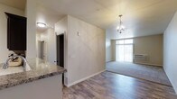 This single-floor residence offers a thoughtfully designed open layout that seamlessly connects the kitchen and living areas. The kitchen features a granite-topped breakfast bar, providing both a functional workspace and a casual dining option. Hardwood-style flooring extends throughout the main living spaces, lending warmth and a cohesive aesthetic to the interior.

A partial wall divides the kitchen from the living area, creating a sense of defined space while maintaining an open feel. The living area is bathed in natural light, thanks to large windows that draw the eye toward the outdoors. An elegant orb-style chandelier serves as a focal point overhead, adding a modern yet classic decorative touch to the space.

The neutral, warm-toned walls throughout complement the light-colored finishes, creating a bright and inviting ambiance. A hallway visible from the main area leads to additional rooms, suggesting a practical and well-organized floor plan. The overall design balances contemporary style with everyday functionality, making it a comfortable and visually appealing living space.