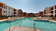 Located at 300 Commerce Ave in LaGrange, Georgia, this outdoor amenity space is designed to deliver a resort-caliber experience within a well-appointed residential community. At its centerpiece sits a large, sparkling swimming pool with crystal-clear turquoise water, framed by an expansive concrete sundeck outfitted with rows of modern lounge chairs and shaded umbrella seating — ideal for relaxing under the open Georgia sky.

The pool area is embraced on multiple sides by multi-story residential buildings featuring a striking mix of warm cedar-toned siding, crisp white vertical board-and-batten panels, and brick accents, creating a cohesive and visually dynamic architectural backdrop. Private balconies on the upper floors overlook the courtyard, adding to the sense of community and connectivity.

A covered pergola-style pavilion anchors one end of the outdoor space, providing a shaded gathering area equipped with ceiling fans, string lights, a mounted television, and recreational amenities including a ping pong table — making it a versatile venue for both relaxation and socializing. Thoughtfully designed landscaping throughout the grounds features ornamental grasses, flowering shrubs, evergreen plantings, and decorative stone beds, adding texture and color to the environment. The overall ambiance is vibrant, welcoming, and meticulously maintained.