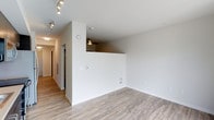 This single-floor space offers a clean, modern aesthetic defined by crisp white walls, light wood-grain flooring, and a thoughtful layout that maximizes openness and flow. The main living area is bright and airy, illuminated by sleek track lighting mounted on the ceiling, which enhances the sense of space throughout. A distinctive architectural feature is the elevated half-wall pass-through that separates the kitchen from the living area, adding visual interest and a split-level dynamic without sacrificing the open feel. The kitchen area above is fitted with its own recessed and track lighting, creating a well-lit, functional workspace. A long corridor extends from the living area, lined with neutral tones and hardwood-style flooring that carries consistently throughout, leading to additional rooms. Floor vents are integrated discreetly into the baseboards, and the walls are clean and neutral, providing a versatile canvas for any interior style. The overall ambiance is contemporary and minimalist, with a fresh, move-in-ready quality that emphasizes practicality and modern design sensibility.