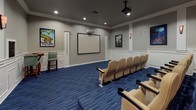 This media center is a purpose-built screening room designed to deliver a true theater experience. The room is anchored by a large, wall-mounted projection screen at the front, paired with a ceiling-mounted projector to ensure crisp, high-quality display. Rows of cushioned, tan theater-style seats with built-in armrests and cup holders are arranged on a tiered platform, providing clear sightlines from every position in the room. The flooring throughout is a vibrant blue patterned carpet that adds a cinematic character to the space. Walls are finished in neutral gray tones and adorned with framed movie posters, lending an authentic movie house atmosphere. White wainscoting panels line the lower portion of the walls, adding a polished, architectural detail. Wall-mounted sconce lighting provides soft ambient illumination to complement the recessed ceiling lights. A small bistro-style table with two bar-height chairs is positioned near the front of the room, offering a casual seating alternative. Dedicated speaker panels are integrated into the walls to support a full surround-sound audio setup. A single entry door provides access to the room, keeping the space self-contained and acoustically focused for an optimal viewing environment.