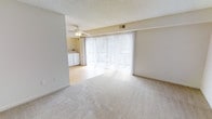 This single-floor space offers a bright and functional layout highlighted by a generous living area with neutral-toned wall-to-wall carpeting that creates a warm, comfortable atmosphere. A prominent set of floor-to-ceiling sliding glass doors dominates one wall, allowing abundant natural light to pour into the room while providing access to an outdoor area. Vertical blinds offer flexible light control throughout the day. A ceiling fan is centrally positioned overhead, contributing to year-round comfort. The textured ceiling adds a classic touch to the interior. An adjacent area features smooth hardwood-style flooring, creating a natural transition between spaces, and includes washer and dryer units, adding everyday convenience. The neutral beige color palette throughout the walls and flooring gives the space a clean, cohesive look that is easy to personalize. Overall, the layout is practical and open, with a calm, well-lit ambiance suited for comfortable everyday living.