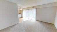 This single-floor space offers a bright and functional layout highlighted by a generous living area with neutral-toned wall-to-wall carpeting that creates a warm, comfortable atmosphere. A prominent set of floor-to-ceiling sliding glass doors dominates one wall, allowing abundant natural light to pour into the room while providing access to an outdoor area. Vertical blinds offer flexible light control throughout the day. A ceiling fan is centrally positioned overhead, contributing to year-round comfort. The textured ceiling adds a classic touch to the interior. An adjacent area features smooth hardwood-style flooring, creating a natural transition between spaces, and includes washer and dryer units, adding everyday convenience. The neutral beige color palette throughout the walls and flooring gives the space a clean, cohesive look that is easy to personalize. Overall, the layout is practical and open, with a calm, well-lit ambiance suited for comfortable everyday living.