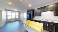 This single-floor space showcases a thoughtfully designed open-concept layout where the kitchen and living area flow seamlessly together. The kitchen is a standout feature, outfitted with dark espresso cabinetry that extends to the ceiling, providing generous storage throughout. A butcher block-topped island anchors the space, offering additional prep area and casual seating potential. Light-toned stone countertops along the perimeter complement the rich cabinetry, while black appliances—including a full-size refrigerator and dishwasher—maintain a cohesive, contemporary aesthetic. Recessed lighting illuminates the kitchen, keeping the space bright and functional. Dark hardwood-style flooring runs throughout, grounding the space with warmth and continuity. The adjacent living area benefits from multiple large windows that invite natural light and provide views of the surrounding neighborhood, creating an airy and open atmosphere. Neutral gray walls throughout offer a versatile backdrop suited to a variety of décor styles. A secondary doorway visible from the main living space suggests additional rooms beyond, adding to the overall functionality of this well-appointed single-floor layout.