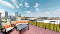 E'laan offers a standout rooftop terrace that serves as a premier outdoor living space with commanding views of the surrounding urban skyline. The terrace is finished with rich, warm-toned composite wood decking that provides a durable and visually appealing surface underfoot. A sleek black metal railing frames the perimeter, ensuring safety while preserving unobstructed sightlines across the cityscape.

A lush green hedge border runs along the railing, introducing a natural element that softens the urban setting and adds vibrant color contrast to the space. The terrace is furnished with a generously sized sectional sofa dressed in cream upholstery and accented with bold striped orange and red throw pillows, paired with a dark wood coffee table — creating a comfortable and stylish outdoor lounge area well-suited for relaxation or social gatherings.

The open-air setting allows for abundant natural light throughout the day, with expansive sky views that shift with the weather and time of day. The surrounding skyline, featuring a mix of high-rise towers and mid-rise buildings, provides a dynamic urban backdrop. Overall, the terrace combines thoughtful design with an inviting atmosphere, making it a distinctive feature of E'laan.