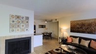 This single-floor residence offers a well-designed open-concept layout that connects the main living areas in a cohesive and functional flow. The living room serves as the heart of the space, anchored by a tile-surround fireplace that adds warmth and a striking architectural focal point. Light hardwood flooring extends throughout, lending a clean, unified look to the interior. A comfortable seating area is complemented by tasteful décor and accent artwork, creating an inviting atmosphere. Adjacent to the living room, a breakfast bar with a dark countertop provides a natural transition into the kitchen, which features dark appliances and ample cabinetry. The dining area, positioned just beyond the kitchen, is illuminated by a ceiling fan with integrated lighting, making it ideal for everyday meals or casual gatherings. Neutral wall tones throughout the space allow the furnishings and decorative accents to stand out, while a textured ceiling adds subtle depth. The overall ambiance is warm and comfortable, with a layout that balances open communal areas with a sense of intimacy, making it a practical and welcoming place to call home.