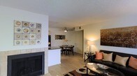 This single-floor residence offers a well-designed open-concept layout that connects the main living areas in a cohesive and functional flow. The living room serves as the heart of the space, anchored by a tile-surround fireplace that adds warmth and a striking architectural focal point. Light hardwood flooring extends throughout, lending a clean, unified look to the interior. A comfortable seating area is complemented by tasteful décor and accent artwork, creating an inviting atmosphere. Adjacent to the living room, a breakfast bar with a dark countertop provides a natural transition into the kitchen, which features dark appliances and ample cabinetry. The dining area, positioned just beyond the kitchen, is illuminated by a ceiling fan with integrated lighting, making it ideal for everyday meals or casual gatherings. Neutral wall tones throughout the space allow the furnishings and decorative accents to stand out, while a textured ceiling adds subtle depth. The overall ambiance is warm and comfortable, with a layout that balances open communal areas with a sense of intimacy, making it a practical and welcoming place to call home.
