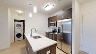 Located at 550 Halstead Ave in Harrison, NY, this well-appointed one-bedroom, one-bathroom residence offers a thoughtfully designed single-floor layout with modern finishes throughout. Light wood flooring flows seamlessly across the living areas, creating a warm, cohesive aesthetic.

The kitchen serves as a functional centerpiece, featuring dark wood cabinetry, a large center island with a white quartz countertop and undermount sink, stainless steel appliances including a side-by-side refrigerator, electric range, and built-in microwave, as well as in-unit washer and dryer conveniently tucked nearby. Pendant lighting and flush-mount fixtures illuminate the space efficiently.

The spacious living area is bright and open, benefiting from large windows that frame lush outdoor greenery, filling the room with natural light. The clean white walls and light flooring contribute to an airy, inviting atmosphere.

The bedroom is generously sized with light wood floors, a dedicated closet, and a large window offering serene outdoor views. A wall-mounted HVAC unit ensures year-round comfort.

The bathroom features a full soaking tub and shower combo, accented by floor-to-ceiling linear tile in soft gray tones and brushed chrome fixtures, offering a clean, contemporary finish throughout.