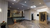 This contemporary lobby space combines modern design elements with functional layout to create a welcoming and professional environment. The floor is finished in a striking herringbone-patterned wood-look tile, adding a sophisticated visual texture that anchors the entire space. Recessed ceiling lighting is complemented by a geometric pendant light fixture, ensuring the area is bright and well-illuminated throughout.

A staircase with sleek glass panel railings serves as a prominent architectural feature, offering both elegance and openness. Adjacent to the staircase, a dark-topped reception or check-in counter provides a practical focal point for visitors. A wall-mounted flat-screen display is positioned nearby, ideal for informational or directional content.

The elevator, framed by smooth, light-toned wall paneling, offers convenient vertical access and contributes to the clean, modern aesthetic. Textured accent walls in neutral tones add depth and visual interest without overwhelming the space. A minimalist bench seat provides a practical resting spot for visitors. The overall ambiance is polished and professional, reflecting a thoughtfully designed commercial or mixed-use environment built to accommodate high foot traffic while maintaining a refined, contemporary atmosphere.