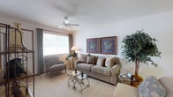 Located at 4111 Wincove Dr in Groveport, Ohio, this single-story home presents a thoughtfully designed interior with a warm and inviting atmosphere. The living room serves as a central gathering space, featuring plush wall-to-wall carpeting that adds a sense of comfort underfoot. A ceiling fan with a light fixture is centrally positioned, providing both air circulation and ambient lighting throughout the room. A large picture window dressed with elegant floor-length drapes allows generous natural light to fill the space while offering views of the surrounding greenery outside. The room is styled with a classic aesthetic, highlighted by a full-sized sofa adorned with decorative pillows, a coordinating accent chair, and an ornate wrought-iron glass-top coffee table that serves as a sophisticated focal point. Two framed botanical art pieces displayed side by side on the wall contribute to the refined, traditional character of the home. Lush indoor plants add a natural, lively touch to the overall ambiance. The neutral color palette of soft whites and warm beiges throughout the space creates a calm and cohesive environment, making this home a comfortable and well-appointed residence in the Groveport community.
