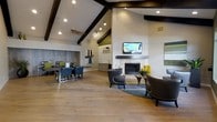 This single-floor space is designed to accommodate both relaxation and social gatherings, showcasing a thoughtfully open layout with distinct functional zones. The most striking architectural feature is the vaulted ceiling accented by bold dark wood beams, which adds dramatic height and visual warmth to the interior. Track lighting and pendant fixtures are strategically placed to illuminate the space effectively.

The lounge area anchors one side of the room, centered around a stone-surround fireplace topped with a wall-mounted flat-screen television. A curved sofa and accent chairs arranged on a circular area rug create an inviting seating arrangement, complemented by yellow accent pieces that add a pop of color. Adjacent to the lounge, a dining area features a large dark wood table surrounded by upholstered chairs, ideal for group gatherings.

A kitchenette or wet bar area is visible in the background, offering functional support for events. The wide-plank hardwood flooring flows seamlessly throughout, unifying each zone. The neutral color palette of warm whites and greiges, paired with green accent walls, gives the space a contemporary yet welcoming ambiance.