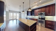 This single-floor residence showcases a thoughtfully designed open-concept layout with distinct living zones connected by rich hardwood flooring. The kitchen serves as the centerpiece, boasting an elongated granite-topped island illuminated by stylish pendant lights, complemented by dark espresso cabinetry — including open display shelving — and a full suite of stainless steel appliances. A light-toned subway tile backsplash adds texture, while track lighting enhances the workspace. The kitchen flows seamlessly toward a living area framed by French doors that invite natural light and offer views to an outdoor balcony.

A dedicated butler's pantry or dry bar area with additional cabinetry and under-cabinet lighting extends the kitchen's functionality. The bedrooms feature neutral carpet, soft wall tones, and windows that provide natural light and exterior views. A spacious walk-in closet with multi-tiered shelving and hanging rods offers generous storage.

The bathrooms are well-appointed, including a primary en-suite featuring floor-to-ceiling gray tile, a glass-enclosed shower with a rain head, a soaking tub, and granite countertops on dark cabinetry. A secondary bathroom mirrors the refined finishes with matching granite surfaces and wood-look tile flooring throughout.