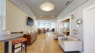 This single-floor space functions as a well-appointed community or leasing clubhouse, offering a comfortable and stylish environment across a generously sized open floor plan. The interior is unified by warm, light-toned wood-look flooring that runs throughout, contributing to a bright and welcoming ambiance.

The main lounge area serves as the central gathering space, furnished with neutral upholstered sofas and chairs arranged to encourage relaxed conversation. A wall-mounted flat-screen television is positioned above a wood credenza, while geometric-patterned wallcovering on an accent wall adds a contemporary design element. Decorative sculptural wall pieces provide additional visual interest along the adjacent corridor.

Toward the rear of the space, a sunlit dining nook features vaulted ceilings with clerestory triangular windows and French doors that open directly to an outdoor pool area, flooding the room with natural light and views of the exterior amenity space. A bistro-height table with upholstered bar stools completes this area.

A separate kitchenette includes white cabinetry, granite countertops, and a peninsula workspace, providing practical utility for hosting or daily use. Recessed lighting and a statement geometric ceiling fixture in the main lounge enhance the overall polished, modern aesthetic of the space.