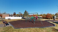 Located at 2828 Egypt Rd in Norristown, Pennsylvania, this residential community presents a well-established setting defined by classic multi-story brick construction with siding accents, teal-trimmed windows, and balconies that add character to each building's facade. The grounds are expansive, offering generous open lawn areas framed by mature evergreen and deciduous trees that provide vibrant seasonal color. Multiple paved parking areas are thoughtfully distributed throughout the property, ensuring convenient access for residents. A dedicated children's play area serves as a central outdoor amenity, equipped with a swing set, a multi-component play structure featuring slides and climbing elements, and rubber mulch safety surfacing contained within a bordered perimeter. A separate, smaller swing area with additional play equipment is also present on the grounds. The community office entrance is attractively landscaped with flowering shrubs and seasonal plantings, and features a covered stairway entry. A covered outdoor kiosk station is positioned near the parking areas for resident use. Wrought iron fencing accents portions of the property boundary, adding a structured, secure feel to the community. The overall environment is open, well-lit with abundant natural light, and thoughtfully maintained, creating an inviting and functional atmosphere for residents.