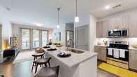This well-designed single-floor residence offers a thoughtful layout that combines modern finishes with functional living spaces. The open-concept kitchen is a standout feature, anchored by a large granite-topped island with an undermount sink, bar seating, and stylish pendant lighting overhead. Light wood-toned cabinetry lines the walls alongside a full suite of stainless steel appliances, including a refrigerator, range, and over-the-range microwave, all set against a clean subway tile backsplash and wood-look flooring. The kitchen flows seamlessly into the living area, which is bathed in natural light from floor-to-ceiling glass doors that open to an outdoor balcony, offering views of the surrounding greenery. The space accommodates comfortable seating and features neutral walls that complement a variety of décor styles. The residence includes multiple bedrooms, each carpeted and filled with natural light from large windows. One bedroom features an ensuite bathroom, while the bathrooms throughout are generously sized, showcasing granite-topped dual vanities, wood-look tile flooring, soaking tubs, and walk-in closets with built-in shelving. The overall aesthetic is contemporary and cohesive, with a warm, neutral palette accented by modern hardware and finishes throughout.