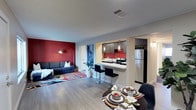 This single-floor space offers a thoughtfully designed open-concept layout that connects the living, dining, and kitchen areas into one cohesive and functional environment. The living area makes a bold visual statement with a striking red accent wall, complemented by a stylish sofa and a colorful area rug that adds warmth and personality to the space. Light wood-toned flooring runs throughout, lending a clean, contemporary feel and creating a sense of continuity between areas. The kitchen is well-appointed with dark cabinetry, a vibrant red subway tile backsplash, stainless steel appliances including a full-size refrigerator, and an electric range. A peninsula with a white countertop extends into the space, providing casual seating with sleek black bar stools and serving as a natural divider between the kitchen and the rest of the living area. Pendant lighting above the peninsula adds both functionality and a modern aesthetic touch. The dining area, positioned adjacent to the kitchen, features a round table set for multiple guests, ideal for everyday meals or entertaining. A passageway leads to additional areas of the unit, suggesting further private spaces beyond the main living zone. Overall, the space exudes a modern, energetic ambiance with carefully chosen design elements.
