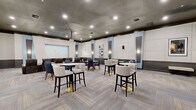 Located at 2801 Biltmore Park Dr in Orlando, FL, this single-floor community amenity space at The Estates at Park Avenue offers a sophisticated and well-appointed environment designed for entertainment and socializing. Guests are welcomed through a marquee-framed entrance, immediately setting a cinematic tone that carries throughout the space.

The main lounge and screening area is expansive, featuring herringbone-patterned carpet, a dark textured ceiling with recessed lighting, and walls adorned with large white acoustic panels accented by light blue pilasters. Multiple seating arrangements include a mix of bar-height tables with upholstered swivel stools on gold bases, standard round tables with navy and cream chairs, and plush built-in banquette seating — all contributing to a flexible, social atmosphere.

A large projection screen anchors the theater-style viewing area, supported by wall-mounted speakers for an immersive experience. Adjacent to the main hall, a dedicated snack bar area features glossy black tile walls, vending machines, a popcorn maker, and gray cabinetry — ideal for movie nights and gatherings.

A bright, welcoming foyer with light wood-look tile flooring and a comfortable seating nook transitions to additional rooms, including a billiards room. The overall design blends modern elegance with functional amenity spaces throughout.