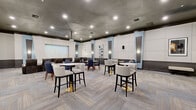 Located at 2801 Biltmore Park Dr in Orlando, FL, this single-floor community amenity space at The Estates at Park Avenue offers a sophisticated and well-appointed environment designed for entertainment and socializing. Guests are welcomed through a marquee-framed entrance, immediately setting a cinematic tone that carries throughout the space.

The main lounge and screening area is expansive, featuring herringbone-patterned carpet, a dark textured ceiling with recessed lighting, and walls adorned with large white acoustic panels accented by light blue pilasters. Multiple seating arrangements include a mix of bar-height tables with upholstered swivel stools on gold bases, standard round tables with navy and cream chairs, and plush built-in banquette seating — all contributing to a flexible, social atmosphere.

A large projection screen anchors the theater-style viewing area, supported by wall-mounted speakers for an immersive experience. Adjacent to the main hall, a dedicated snack bar area features glossy black tile walls, vending machines, a popcorn maker, and gray cabinetry — ideal for movie nights and gatherings.

A bright, welcoming foyer with light wood-look tile flooring and a comfortable seating nook transitions to additional rooms, including a billiards room. The overall design blends modern elegance with functional amenity spaces throughout.