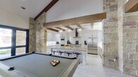 This impressive single-floor amenity space showcases a sophisticated blend of rustic and modern design. The grand entrance features bold double black doors framed by natural limestone columns, with a vaulted beamed ceiling and a dramatic globe pendant chandelier setting the tone for the entire space.

The expansive main lounge area commands attention with its soaring ceilings adorned by exposed dark wood beams and striking Sputnik-style chandeliers. Towering limestone columns define the open floor plan while large, dark-framed windows and glass doors flood the interior with natural light and offer views to the outdoors. A full-sized billiards table anchors one side of the room, complemented by generous sectional seating arranged over patterned area rugs. A floor-to-ceiling stone fireplace with a rustic wood mantel serves as a dramatic focal point along one wall.

The fully equipped kitchen is a standout feature, offering professional-grade stainless steel appliances including a multi-burner gas range with a sleek range hood, a French door refrigerator, and a large island with bar seating. White shaker cabinetry, quartz countertops, a marble-style backsplash, and geometric pendant lighting complete the polished, functional aesthetic throughout the kitchen. Light wood-look flooring flows seamlessly across the entire space, tying together every area cohesively.