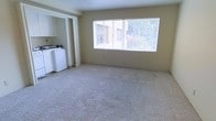 This single-floor space offers a straightforward, functional layout designed for comfortable everyday living. The main living area is finished with wall-to-wall carpet in a neutral tone, providing a soft underfoot feel and a clean, cohesive look throughout. A large picture window dominates one wall, allowing ample natural light to fill the room while offering views of the surrounding exterior. The bright, open feel of the space makes it well-suited for a variety of furniture arrangements.

Adjacent to the living area, an open pass-through connects to what appears to be a combined kitchen and laundry space. White cabinetry lines the upper walls of this area, providing functional storage. White appliances, including what appears to be a refrigerator and washer, are visible, indicating that both kitchen and laundry facilities are conveniently located within the same footprint.

The overall color palette is neutral throughout, with beige walls complementing the light carpet. The single-floor layout ensures easy accessibility, while the open connection between the living and utility areas contributes to an efficient use of space. The design is clean and practical, offering a blank canvas ready for personalization.