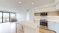 Located at 747 Hampden Ave in Saint Paul, MN, this modern 2-bedroom, 2-bathroom residence offers a thoughtfully designed single-floor layout with clean lines and a bright, contemporary aesthetic throughout. The open-concept kitchen and living area serves as the heart of the unit, featuring light hardwood flooring, recessed lighting, and a central island with a white quartz countertop — ideal for dining or meal prep. The kitchen is outfitted with sleek white cabinetry accented by warm wood-toned panels, under-cabinet lighting, and a full suite of stainless steel appliances including a range, microwave, dishwasher, and refrigerator. The living area extends to a sliding glass door that opens to a private balcony, offering views of the surrounding Saint Paul neighborhood. The spacious primary bedroom features plush carpet, generous proportions, and a large window that fills the room with natural light. The second bedroom is similarly carpeted and well-sized. Both bathrooms are finished with floating white vanities, large mirrors, brushed nickel fixtures, and hardwood-style flooring. The unit's clean white walls and modern finishes create a cohesive, move-in-ready living environment.