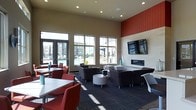 This single-floor community clubroom is designed for both socializing and relaxation, offering a well-appointed gathering space with a thoughtful layout and contemporary finishes. The room is defined by soaring ceilings and a bold accent wall in deep red, which adds a striking visual contrast to the neutral beige tones throughout the space. Recessed lighting illuminates the interior, while a row of large windows floods the room with abundant natural light and provides unobstructed views of an outdoor pool and surrounding residential buildings.

The space is organized into multiple functional zones. A dining area along the perimeter features white rectangular tables paired with vibrant red chairs, ideal for casual meals or group gatherings. A central lounge area offers plush dark upholstered seating arranged around low round tables, creating an inviting setting for conversation. Adjacent to this, a sleek wall-mounted linear fireplace serves as a focal point alongside a large flat-screen television, perfect for entertainment.

Dark patterned carpeting grounds the space and ties together the various seating arrangements, while the overall color palette of red, charcoal, and white lends a polished, modern aesthetic. The combination of natural light, varied seating options, and quality finishes makes this a versatile and welcoming communal space.