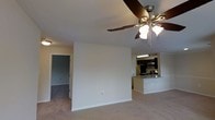 This single-floor residential space offers a clean, open-concept layout designed for comfortable everyday living. The main living area is finished with soft wall-to-wall carpeting and crisp white walls, creating a bright and welcoming atmosphere. A wood-toned ceiling fan with integrated lighting serves as a functional focal point overhead, providing both air circulation and ambient illumination throughout the space.

The kitchen is positioned in an open-plan arrangement adjacent to the living area, separated by a peninsula-style breakfast bar with a light-toned countertop that doubles as a casual dining surface. The kitchen is equipped with dark wood cabinetry, stainless steel appliances including a microwave and refrigerator, and recessed overhead lighting, giving it a contemporary and practical feel. The contrast between the dark cabinetry and light countertops adds visual interest to the cooking space.

A doorway visible from the living area leads to an additional room, suggesting a bedroom or ancillary space finished with matching carpeted flooring. The neutral color palette used throughout the unit — from the walls to the flooring — lends itself well to a variety of furnishing styles. Overall, the space presents a functional and well-appointed interior suited for residential living.