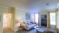 This single-story space centers around a well-appointed living area designed with a calm, neutral aesthetic. The living room features plush carpeted flooring and light-toned walls accented by crown molding, lending a clean and polished finish to the space. A large, comfortable sofa serves as the focal point, adorned with decorative throw pillows in muted patterns, complemented by a sleek coffee table with hairpin legs. Ambient lighting is provided by stylish table lamps, while a framed piece of wall art adds a subtle decorative touch. A window dressed with sheer curtains allows natural light to filter softly into the room, contributing to its warm and inviting atmosphere. An open archway connects the living area to an adjacent room, which is partially visible and features a bookcase or media storage unit, suggesting a functional secondary space. The overall layout feels open and cohesive, with thoughtful furniture placement that maximizes the sense of space while maintaining a comfortable and livable environment throughout.