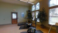 This fitness room offers a practical and welcoming environment for exercise, housed within a single-floor facility. The space is brightened by a pair of elegant arched windows adorned with wood-framed blinds, allowing generous natural light to fill the room while providing views of the surrounding greenery outside. The walls are painted in a soft sage green, lending a calm and refreshing ambiance that complements an active setting.

The room is equipped with a selection of cardio machines, including a treadmill, an elliptical trainer, and an upright stationary bike, each placed on protective rubber floor mats. A mounted flat-screen television is positioned conveniently for viewing during workouts, while a heart rate reference chart is displayed on the wall for easy consultation. Decorative potted trees are thoughtfully placed throughout the space, adding a touch of nature to the interior.

Overhead fluorescent lighting supplements the natural light, ensuring the room remains well-lit at all times. A classic analog wall clock keeps exercisers on schedule, and the clearly marked exit door with a wood finish provides safe and easy access. The overall atmosphere is clean, organized, and functional, making this fitness room a convenient and comfortable space for daily exercise routines.