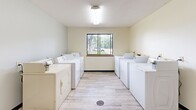This shared laundry facility at 1804 University Dr SE in Saint Cloud, Minnesota, offers a clean, functional, and well-organized space designed for convenient communal use. The room is finished with light-toned vinyl plank flooring throughout, contributing to a bright and easy-to-maintain environment. Overhead fluorescent lighting ensures the space is well-lit at all times.

The main laundry area features rows of full-size top-loading washers and dryers lined along both sides of the room, providing ample capacity for multiple users simultaneously. A central window along the back wall allows natural light to filter in and offers a view of the surrounding outdoor greenery, enhancing the overall atmosphere of the space.

A dedicated utility sink station is positioned along one wall, offering a practical resource for hand-washing or pre-treating garments. A small table provides a convenient surface for folding or sorting laundry. The room also includes a waste bin for general use and is accessible through a solid wood-panel entry door equipped with an automatic door closer.

The walls are finished in a soft, neutral tone, and the textured ceiling completes the clean, utilitarian aesthetic. Overall, this laundry facility is a well-equipped, practical space that serves the needs of its users efficiently and comfortably.