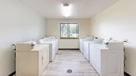 This shared laundry facility at 1804 University Dr SE in Saint Cloud, Minnesota, offers a clean, functional, and well-organized space designed for convenient communal use. The room is finished with light-toned vinyl plank flooring throughout, contributing to a bright and easy-to-maintain environment. Overhead fluorescent lighting ensures the space is well-lit at all times.

The main laundry area features rows of full-size top-loading washers and dryers lined along both sides of the room, providing ample capacity for multiple users simultaneously. A central window along the back wall allows natural light to filter in and offers a view of the surrounding outdoor greenery, enhancing the overall atmosphere of the space.

A dedicated utility sink station is positioned along one wall, offering a practical resource for hand-washing or pre-treating garments. A small table provides a convenient surface for folding or sorting laundry. The room also includes a waste bin for general use and is accessible through a solid wood-panel entry door equipped with an automatic door closer.

The walls are finished in a soft, neutral tone, and the textured ceiling completes the clean, utilitarian aesthetic. Overall, this laundry facility is a well-equipped, practical space that serves the needs of its users efficiently and comfortably.