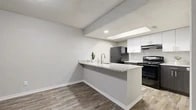 This single-floor residence offers a cohesive, modern aesthetic anchored by wood-look flooring that runs consistently throughout every room. The living room serves as a welcoming centerpiece, featuring a white brick fireplace, natural light from large windows with blinds, and a ceiling fan overhead. An open-concept layout connects the living area to the kitchen, which is defined by a peninsula counter with granite-look surfaces and an integrated sink—ideal for casual dining or food preparation. The kitchen itself is well-equipped with dark lower cabinetry, white upper cabinets, a range with electric cooktop, and a full-size refrigerator, all topped with matching stone-look countertops. A skylight above the kitchen area introduces additional natural light. The bedroom features a walk-in closet, two additional doors for access and storage, and ample floor space. A secondary bedroom is bright with a window and ceiling fan. The bathroom includes a dark vanity cabinet with stone countertop and wood-look flooring. Textured ceilings with recessed lighting appear throughout, and the neutral gray wall palette creates a clean, consistent look across the entire space.
