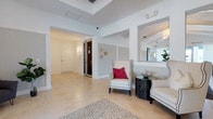 This single-floor space presents a clean, contemporary aesthetic defined by crisp white walls, light-toned large-format tile flooring, and thoughtfully designed architectural details. The entry and living area immediately draw attention with a coffered tray ceiling that adds depth and dimension to the interior, while recessed and wall-mounted lighting ensures a warm, well-lit ambiance throughout.

The seating area serves as a focal point, featuring a classic high-back wingchair upholstered in cream fabric with contrasting piping and accented by a vibrant fuchsia pillow. It is paired with a sleek, dark circular side table and a complementary sofa, creating a refined and inviting arrangement. A large mirror positioned in an arched niche reflects light and visually expands the space, contributing to its airy feel.

A patterned area rug anchors the seating area, adding texture to the smooth tile floor. The hallway leading further into the space is flanked by white panel doors and features a dark wood door, providing contrast against the predominantly light palette. Wall sconces along the corridor offer subtle, decorative illumination. Decorative wall accents and a potted plant introduce organic touches that soften the structured, modern interior design. Overall, the space exudes a polished, sophisticated atmosphere.