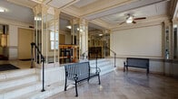 This single-floor space showcases a stately lobby with rich classical architectural detailing throughout. The entrance area features polished marble tile flooring in a large-format pattern, complemented by marble wainscoting that lines the lower walls. Ornate plaster crown molding and coffered ceiling details with gold accent trim lend a formal, refined character to the space. Decorative Corinthian-style columns frame a raised interior platform area, accessible via marble steps fitted with black iron handrails. Mirrored wall panels amplify the sense of depth and grandeur throughout the lobby. Ceiling fans with integrated light fixtures provide functional overhead lighting, while flush-mount fixtures illuminate the connecting corridor. Black iron benches provide seating within the lobby, maintaining the classical aesthetic. A long interior hallway lined with mailboxes extends from the main lobby, with dark-toned carpeting contrasting the marble of the entrance. Large windows along one wall admit abundant natural light, offering views of the surrounding cityscape. Entry doors with glass panels connect the lobby to the exterior, further enhancing brightness. The overall ambiance is one of historic elegance and architectural distinction, characteristic of early 20th-century institutional or residential building design.