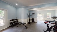 This fitness center in Horn Lake, Mississippi, offers a thoughtfully designed workout facility spread across a single floor, combining functional exercise spaces with refined architectural charm. The interior is finished with soft blue walls, crisp white wainscoting, and elegant crown molding throughout, creating an inviting and motivating environment for exercise.

The facility is organized into distinct workout zones. The cardio area houses two treadmills positioned near windows that overlook outdoor greenery, allowing natural light to fill the space. Additional cardio equipment includes stationary spin bikes and a rowing machine, providing a comprehensive range of aerobic options.

The strength training zone features a multi-function cable machine and a full dumbbell rack with an adjustable weight bench, accommodating a wide variety of resistance exercises. A dedicated wall-mounted rack organizes resistance bands, suspension trainers, and jump ropes for functional fitness workouts.

Protective rubber interlocking floor tiles cover the workout areas, providing both safety and durability underfoot. French doors and large windows connect the space visually to the outdoors while ensuring ample natural light. Recessed lighting supplements illumination throughout. The layout flows openly between zones, making navigation intuitive and efficient. Overall, the fitness center delivers a clean, polished, and fully functional environment suitable for a broad range of training styles and fitness levels.