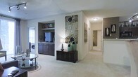 This single-floor residence offers a thoughtfully arranged open layout that balances functionality with aesthetic appeal. The main living area is finished with plush wall-to-wall carpeting in a neutral tone, creating a warm and welcoming atmosphere throughout. A standout feature of the living space is the built-in entertainment nook, which includes dark wood cabinetry with multiple drawers and an open shelf above, providing ample storage and display space for media equipment and personal décor.

The neutral color palette — soft grays and warm whites — lends the space a clean, contemporary feel. A decorative console table with dark wood detailing serves as a stylish focal point, accented by teal ceramic vases and a modern table lamp that add character to the interior. Large-scale wall art panels above the console further contribute to the curated ambiance.

A well-lit hallway extends from the main living area, connecting to additional rooms within the residence. Along the hallway, glimpses of what appears to be a bathroom with light-toned countertops are visible, suggesting well-appointed secondary spaces. Natural light filters into the living area through a window, enhancing the overall brightness of the home. The layout flows intuitively, making the space feel both organized and comfortably livable.