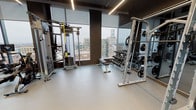 This contemporary fitness center is designed to deliver a comprehensive workout experience across two distinct zones. The main gym area is outfitted with an impressive array of professional-grade equipment, including Life Fitness cable machines, a TRX suspension training station, plate-loaded power racks, a full dumbbell rack, adjustable benches, treadmills, and a rowing machine. Large floor-to-ceiling windows line the space, flooding the gym with natural light while offering panoramic views of the surrounding urban skyline. Full-length mirrors are strategically placed throughout, enhancing the sense of openness and allowing users to monitor their form. Linear LED lighting runs along the dark ceiling, contributing to a sleek, modern aesthetic, while durable light-gray flooring ties the space together. A dedicated cycling studio adjoins the main floor, featuring multiple stationary bikes, wall-mounted televisions, floor-to-ceiling mirrors, and hardwood-style flooring — creating an immersive environment ideal for indoor cycling sessions. Glass doors connect the fitness center to an outdoor terrace, providing a seamless transition between indoor and outdoor spaces. The overall design balances functionality with a refined, upscale ambiance.