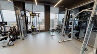 This contemporary fitness center is designed to deliver a comprehensive workout experience across two distinct zones. The main gym area is outfitted with an impressive array of professional-grade equipment, including Life Fitness cable machines, a TRX suspension training station, plate-loaded power racks, a full dumbbell rack, adjustable benches, treadmills, and a rowing machine. Large floor-to-ceiling windows line the space, flooding the gym with natural light while offering panoramic views of the surrounding urban skyline. Full-length mirrors are strategically placed throughout, enhancing the sense of openness and allowing users to monitor their form. Linear LED lighting runs along the dark ceiling, contributing to a sleek, modern aesthetic, while durable light-gray flooring ties the space together. A dedicated cycling studio adjoins the main floor, featuring multiple stationary bikes, wall-mounted televisions, floor-to-ceiling mirrors, and hardwood-style flooring — creating an immersive environment ideal for indoor cycling sessions. Glass doors connect the fitness center to an outdoor terrace, providing a seamless transition between indoor and outdoor spaces. The overall design balances functionality with a refined, upscale ambiance.