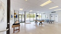 Located at 29106 Lankford Hwy in Cape Charles, Virginia, this single-floor recreational space offers a generously sized open-plan layout designed for entertainment and social gatherings. The interior is defined by gleaming polished terrazzo floors that reflect natural light throughout the room, contributing to a bright and airy atmosphere.

A striking wall of floor-to-ceiling windows runs along the front of the space, flooding the interior with abundant natural light and providing clear views of the surrounding greenery and streetscape. The drop-panel ceiling with recessed lighting ensures the space remains well-lit at all hours.

The layout accommodates a diverse selection of recreational games, including a pool table, ping-pong table, shuffleboard table, cornhole boards, and a putting green strip along the window line, making it a well-equipped venue for competitive and casual play alike. A lounge area with seating, bistro-style high-top tables, and a wall-mounted flat-screen television provides a comfortable space to relax between activities.

Additional features include interior doors leading to separate rooms, wall art that adds a decorative touch, and roller shades for light control. The open, flexible floor plan and bright, clean aesthetic make this space well-suited for events, group recreation, or community use.