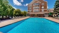 Located at 3021 Maple Valley Dr in Madison, Wisconsin, this outdoor amenity space centers around a generously sized swimming pool with crystal-clear blue water. The pool is equipped with lane dividers, making it suitable for both recreational swimming and lap swimming. Surrounding the pool is a spacious concrete sun deck lined with rows of lounge chairs, providing ample space for relaxation and sunbathing. The area is enclosed by a metal fence, ensuring privacy and safety, while a backdrop of mature trees adds a natural, park-like ambiance to the setting. Floodlights are positioned throughout the deck, allowing for evening use of the pool area. Direct access to the adjacent multi-story building is provided through a covered entrance with glass-paneled doors, seamlessly connecting the outdoor pool area to the interior facilities. The building itself features a classic design with warm brown siding, brick accents, and multiple windows that overlook the pool area. Under open skies and surrounded by lush greenery, this outdoor pool space offers a welcoming and functional retreat for residents or guests seeking both leisure and active recreation.