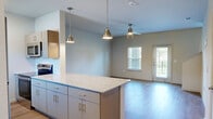 Located at 2902 Pat Booker Rd in Universal City, Texas, this two-story space offers a thoughtfully organized layout across two floors. The ground floor hosts the main living and kitchen area, where warm light wood-style flooring flows throughout. The kitchen is well-appointed with granite countertops, contemporary gray cabinetry, stainless steel appliances including a range, microwave, dishwasher, and refrigerator, plus a central island with pendant lighting. The open living area features a ceiling fan, a glass door, and windows that allow natural light to fill the space. A staircase connects to the upper floor, which includes a hallway, a walk-in closet with wire shelving, and access to the bathrooms. The bathrooms are finished with matching granite vanity counters, gray cabinetry, wood-style flooring, and wall-mounted storage. Neutral tones and clean lines define the overall aesthetic throughout. A balcony off the upper level overlooks a landscaped green area, offering views of the surrounding Universal City neighborhood. The consistent use of light-toned walls and flooring creates a bright, cohesive ambiance across the entire space.