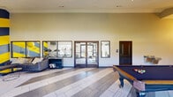 Located at 5151 Highland Rd in Baton Rouge, Louisiana, this single-floor community clubhouse delivers a dynamic and well-appointed gathering space designed for both recreation and productivity. The open-plan layout is anchored by soaring vaulted ceilings and striking large-format tile flooring laid in a bold diagonal pattern, lending a sense of expansive, airy openness throughout.

The lounge area is the visual centerpiece of the space, featuring a dramatic accent wall with bold yellow and charcoal diagonal stripes, a wall-mounted flat-screen TV, and a comfortable sectional sofa arrangement set over a plush area rug. A sleek geometric glass coffee table completes the modern aesthetic. Adjacent to the lounge, a dedicated business center offers workstations with desktop computers positioned beneath large windows that flood the area with natural light and provide views of the surrounding property.

The game zone includes a full-size billiards table and a dartboard mounted to the wall, creating an engaging entertainment area. A wet bar with counter seating is seamlessly integrated nearby, adding convenience for social gatherings. Large glass doors along one wall connect visually to an adjacent fitness facility, maintaining an open and connected feel. Track lighting throughout ensures the entire space is well-lit, while multiple windows and glass doors invite ample natural light from all sides.